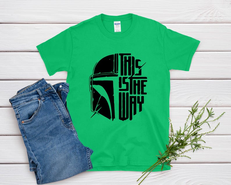 May include: A green t-shirt with a black graphic of a helmet and the text "THIS IS THE WAY." The shirt is paired with blue jeans. A sprig of greenery rests on the wooden surface.