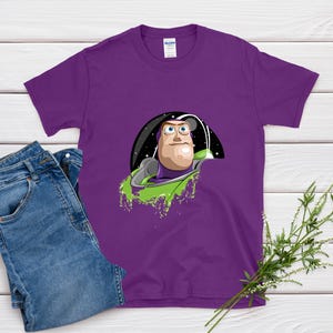 May include: A purple t-shirt featuring a graphic of a cartoon character in a space-themed setting. The character has a light skin tone, a green and purple space suit, and a determined expression. The shirt is paired with blue jeans.
