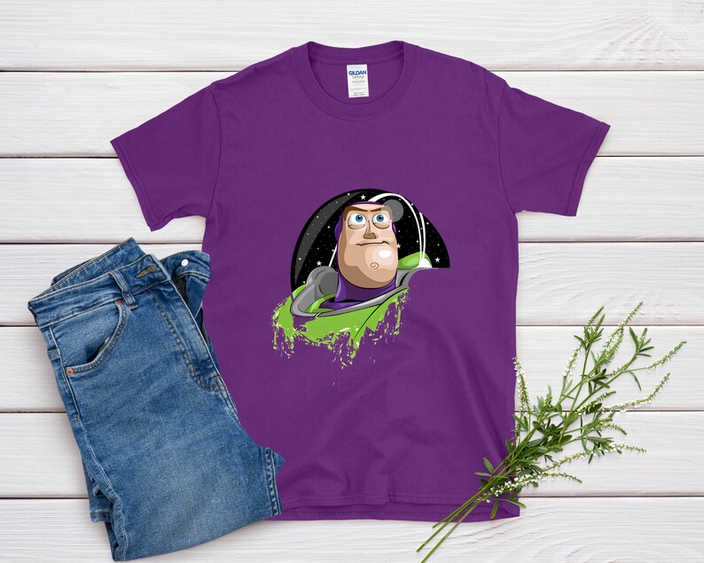 May include: A purple t-shirt featuring a graphic of a cartoon character in a space-themed setting. The character has a light skin tone, a green and purple space suit, and a determined expression. The shirt is paired with blue jeans.