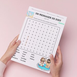 Peut inclure: Une feuille de papier blanche avec un jeu de mots cachés intitulé "UN MESSAGER DE DIEU". Le jeu comprend des mots comme "ESPRIT" et "SAINT". La feuille est tenue par deux mains sur un fond rose.