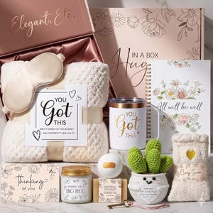 May include: A gift box with a variety of items, including a blanket, eye mask, mug, candle, soap, and a small cactus. The box and items have a soft, neutral color palette with floral accents and the text "You Got This".