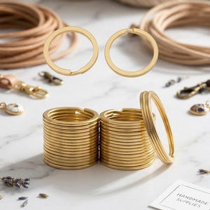 May include: A collection of gold-toned metal split key rings arranged in two neat stacks, with two additional rings floating above. A small white card with the printed text HANDMADE SUPPLIES is visible in the lower right corner of the frame.
