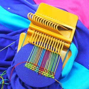 May include: A yellow plastic weaving loom with a blue circular fabric base. The loom has multiple hooks and is partially threaded with colorful yarn in red, yellow, green, and blue. The background is a mix of blue, pink, and turquoise fabric.