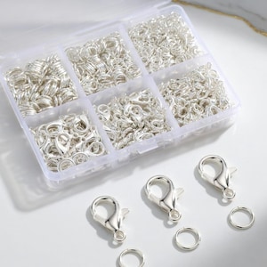 May include: A clear plastic organizer filled with silver-colored jewelry findings. The organizer contains various sizes of jump rings, lobster clasps, and split rings. Three lobster clasps and two jump rings are placed outside the organizer.