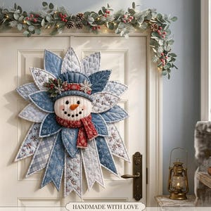 May include: A quilted fabric snowman wall hanging featuring a central snowman face with an orange carrot nose, wearing a blue hat and a red scarf, surrounded by layered, petal-like fabric segments in shades of blue, white, and grey patterns.