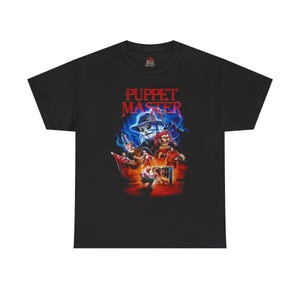 Peut inclure: T-shirt noir avec un graphisme coloré du film "Puppet Master". Le motif présente des personnages de marionnettes, dont un personnage au visage de crâne et un bouffon. Le titre "Puppet Master" est en rouge et bleu.