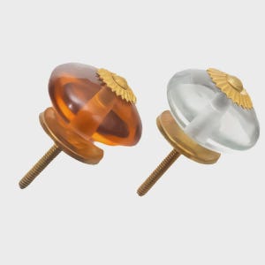 May include: Two decorative cabinet knobs. One is amber-colored, the other is clear. Both have a gold-colored, sunburst-shaped accent and a brass-colored base and screw. These knobs are designed for furniture.