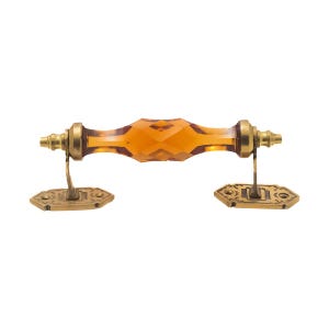 May include: An amber-colored, faceted glass door pull with brass hardware. The handle has a geometric design and is mounted on decorative brass plates. The hardware is a vintage style.