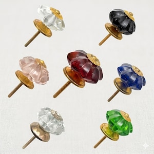 May include: A collection of seven decorative cabinet knobs featuring faceted, pumpkin-shaped glass heads in clear, black, pink, dark red, blue, and green. Each knob has a brass-toned metal base and a threaded screw post for installation.