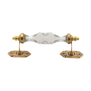 May include: A clear crystal door pull with a faceted design, set between brass-colored hardware. The hardware includes decorative, hexagonal backplates and connecting posts. The door pull is designed for furniture or cabinetry.