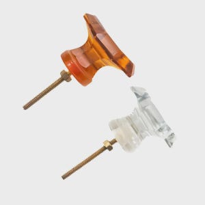 May include: Two decorative cabinet knobs. One is amber-colored, the other is clear. Both knobs have a faceted design and are mounted on threaded rods with nuts. The knobs are likely for furniture or other decorative applications.