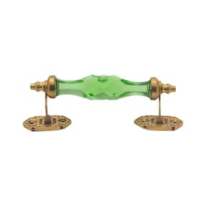May include: A decorative door pull with a faceted, emerald green glass handle. The handle is flanked by antique brass-colored metal hardware. The hardware includes decorative backplates and connecting posts.