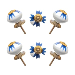 May include: Six decorative cabinet knobs. Each knob features a white ceramic base with blue floral accents and a gold-colored metal top. The knobs have a brass-colored metal screw for installation.