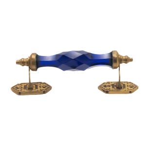 May include: A decorative door handle with a faceted, deep blue glass center and brass-colored metal hardware. The handle is horizontal, with ornate rectangular base plates on each end.