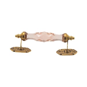 May include: A decorative cabinet pull with a faceted, light pink glass handle. The handle is flanked by ornate, gold-colored metal hardware and mounting plates. The hardware has a vintage aesthetic.