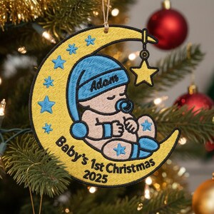 May include: Embroidered Christmas ornament featuring a baby sleeping on a crescent moon. The ornament is yellow with blue accents, stars, and the text "Baby's 1st Christmas 2025". The baby wears a blue cap with the name "Adam".