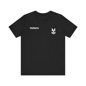 May include: A black short-sleeved t-shirt featuring the white text TARKOV printed on the left chest area, accompanied by a small white graphic emblem of a bird with wings spread above a globe and a banner.