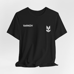 Könnte beinhalten: Schwarzes T-Shirt mit dem weißen Aufdruck "TARKOV" auf der linken Brust. Auf der rechten Brust befindet sich ein weißes Emblem. Das T-Shirt hat kurze Ärmel und einen Rundhalsausschnitt.