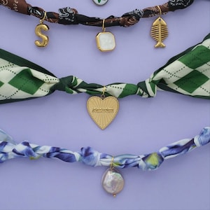 Scarf necklaces with floral and geometric patterns. Scarves have charms attached to them that include the letter "S", pears, gold heart, and a fish.