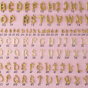 An initial letter collection of gold plated stainless steel charms. Charms include four different styles of initial letters. Styles include big bubble letters, small bubbles, plain, and bamboo letters. Charms are laid out on a pink background with identifying numbers.