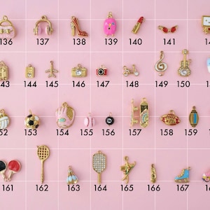 A collection of gold plated stainless steel charms. Charms include purse, headphones, high heels, luggage, travel theme, music instruments, and sports theme like football and pickleball. Charms are laid out on a pink background with identifying numbers.