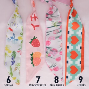 May include: Four colorful fabric scarves with different designs. One is white with a floral pattern, another is pink with strawberries, a third is white with pink tulips, and the last is teal with orange hearts. Each scarf has a number and a descriptive word.