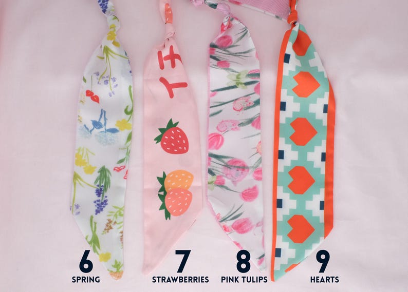 May include: Four colorful fabric scarves with different designs. One is white with a floral pattern, another is pink with strawberries, a third is white with pink tulips, and the last is teal with orange hearts. Each scarf has a number and a descriptive word.