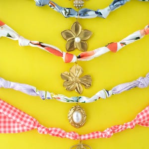May include: A collection of fabric scarf necklaces with various gold-toned pendants, including a bee, flowers, and an oval with a pearl. The fabric strands are in a variety of colors and patterns, set against a bright yellow background.