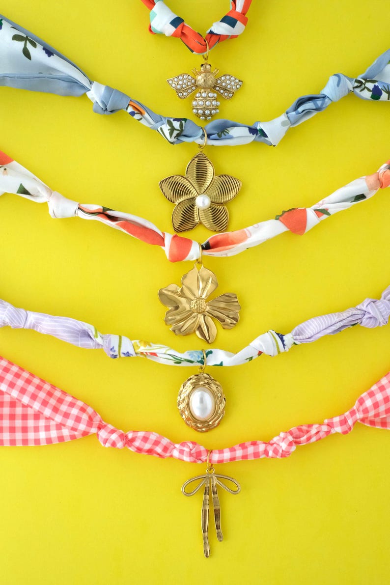 May include: A collection of fabric scarf necklaces with various gold-toned pendants, including a bee, flowers, and an oval with a pearl. The fabric strands are in a variety of colors and patterns, set against a bright yellow background.