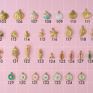A collection of gold plated stainless steel charms in ocean theme. Charms include shells, pearls, anchors, fish, dolphin, starfish, and seahorses. Charms are laid out on a pink background with identifying numbers.