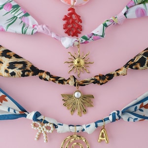 May include: An assortment of colorful scarf necklaces with decorative charms. The headbands feature floral, leopard print, and patterned designs. Charms include a coral, sun, fan, bow, spiral, and the letter "A". The background is a solid pink.