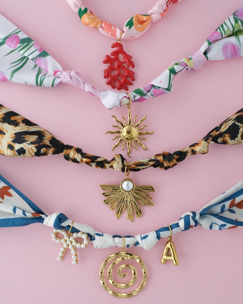 May include: An assortment of colorful scarf necklaces with decorative charms. The headbands feature floral, leopard print, and patterned designs. Charms include a coral, sun, fan, bow, spiral, and the letter "A". The background is a solid pink.