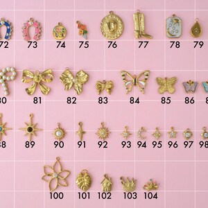 A collection of gold plated stainless steel charms. Charms include lucky charms, cowboy boot and hat, bows, butterflies, stars, moon, monstera leaf, and king crowns. Charms are laid out on a pink background with identifying numbers.