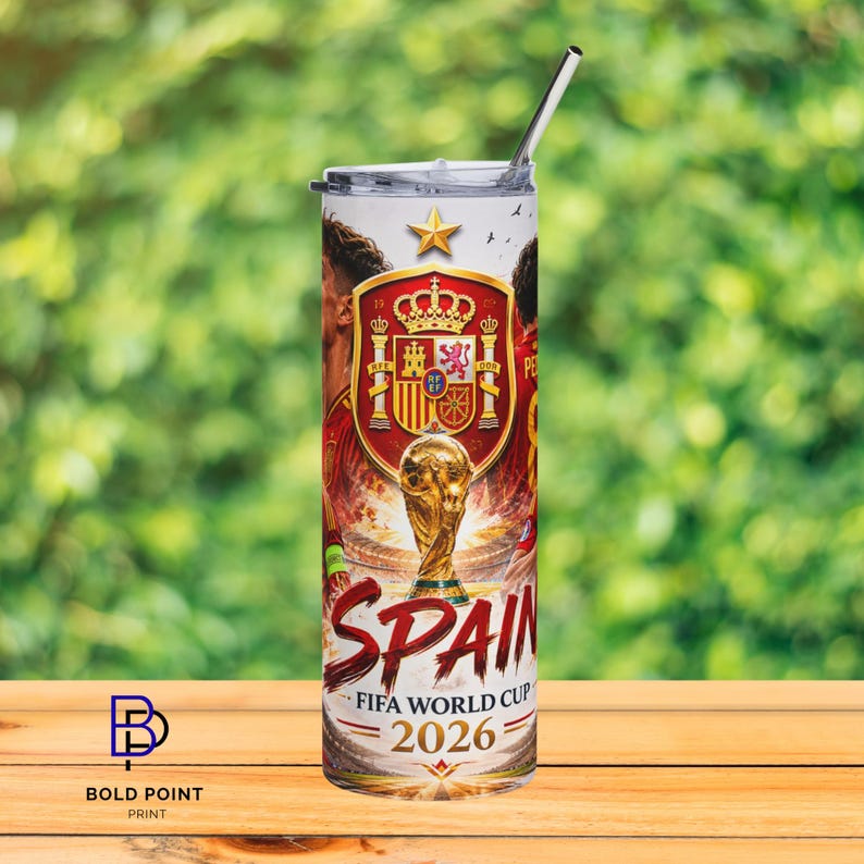 K&ouml;nnte beinhalten: Ein zylindrischer Becher mit Metallstrohhalm, der mit dem spanischen Staatswappen, einem goldenen Pokal und dem Text SPAIN FIFA WORLD CUP 2026 bedruckt ist. Das Design zeigt rote und gelbe Akzente sowie einen Stern &uuml;ber dem Wappen.