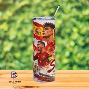 K&ouml;nnte beinhalten: Ein zylindrischer Edelstahlbecher mit einem Grafikdruck der spanischen Fu&szlig;ballnationalmannschaft, der zwei Spieler in roten Trikots, die spanische Flagge und ein Stadion zeigt. Der Becher hat einen transparenten Deckel und einen Metallstrohhalm.
