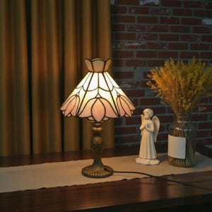 May include: A stained glass table lamp with a floral design. The lamp has a cream-colored shade with pink and white accents, and a bronze-colored base. The lamp is on a wooden table with a neutral-colored runner. A small angel statue and a vase of yellow flowers are also on the table.