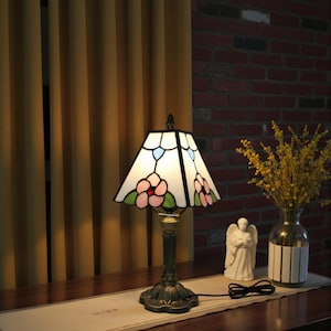 May include: A stained glass table lamp with a floral design. The lamp features a white shade with pink flowers, green leaves, and blue accents. The base is bronze-colored and ornate.