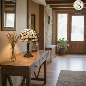 Puede incluir: Una lámpara de mesa decorativa con una pantalla de vidrieras con un diseño de mosaico de piezas de vidrio beige, marrón y coloridas. La lámpara se asienta sobre una base de metal oscuro y se coloca sobre una consola de madera. Un jarrón de cristal transparente con flores secas a la izquierda.