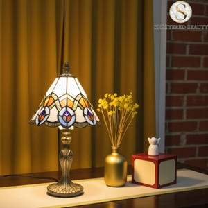 Puede incluir: Una lámpara de mesa de vidrieras con una pantalla blanca y multicolor. La lámpara tiene una base de color bronce y un diseño decorativo. La lámpara está sobre una mesa con un jarrón dorado y una caja roja.