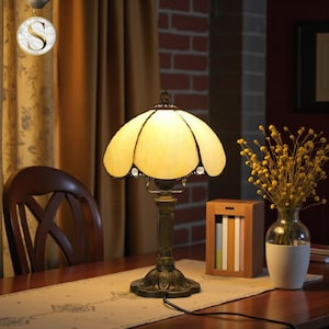 Puede incluir: Una lámpara de mesa decorativa con una pantalla de vidriera amarilla con un diseño floral. La lámpara tiene una base y un tallo de color bronce. La lámpara está sobre una mesa de madera con un camino de mesa floral.