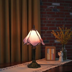 May include: A decorative table lamp with a pink and white floral-shaped shade. The lamp has a bronze-coloured base and stem. The lamp is on a table with a decorative runner. A brick wall and curtains are in the background.