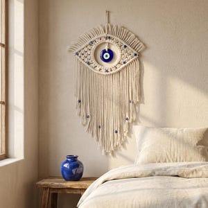 May include: Cream-colored macrame wall hanging in an eye shape, featuring a blue and white glass eye in the center. The macrame has a fringe design and is adorned with small blue beads. A blue vase sits on a wooden table.