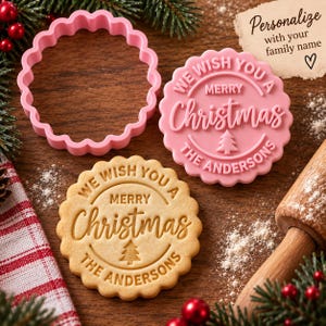 Op de afbeelding: Een roze, geschulpte koekjesvorm en een bijpassende roze stempel met de tekst WE WISH YOU A MERRY CHRISTMAS THE ANDERSONS en een klein kerstboomicoon. Daarnaast ligt een gebakken koekje met hetzelfde reliëf en een houten deegroller.
