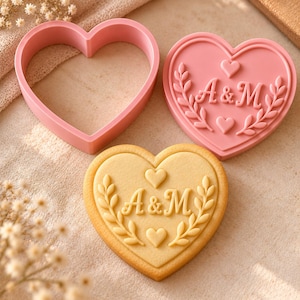 Puede incluir: Un cortador de galletas rosa en forma de corazón, un sello rosa a juego en forma de corazón con las letras A&M rodeadas de ramas de hojas y corazones pequeños, y una galleta beige en forma de corazón con el mismo diseño A&M en relieve.