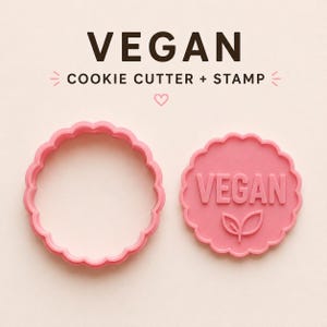 Könnte beinhalten: Ein rosafarbener, gewellter Keksausstecher und ein passender runder Stempel mit dem geprägten Text VEGAN und einem kleinen Blattsymbol. Der Text VEGAN COOKIE CUTTER + STAMP steht in dunkelbraunen Buchstaben mit einem kleinen Herz darüber.