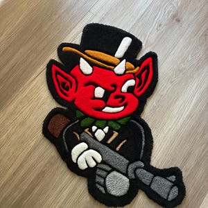 May include: A tufted rug shaped like a cartoon devil character with red skin, white horns, and a black top hat. The character wears a black suit with a green collar and holds a grey object. The design features bold black outlines and vibrant colors.