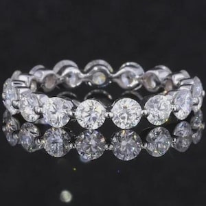 May include: A silver eternity ring with round, clear gemstones set closely together. The ring is highly reflective, with a dark background that enhances the sparkle of the stones. The ring's design is classic and elegant.