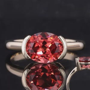 May include: A close-up of a ring with a large, oval-cut, reddish-orange gemstone set in a silver band. The ring is displayed on a reflective surface, with a second ring visible in the background.