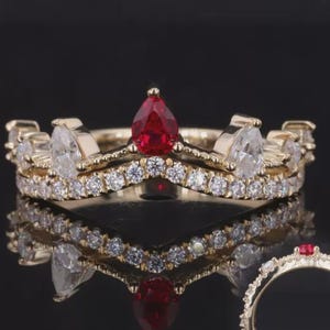 Op de afbeelding: Een goudkleurige ring met een kroonontwerp. De ring heeft een peervormige rode edelsteen in het midden en kleinere witte edelstenen. De ring wordt weerspiegeld op een zwarte ondergrond.