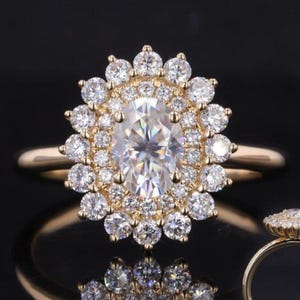 Pode incluir: Um anel de ouro com uma grande pedra central de diamante de corte oval rodeada por um halo de diamantes menores. O anel é montado em uma banda de ouro, e uma versão menor do anel é visível à direita. O anel é exibido em uma superfície preta.